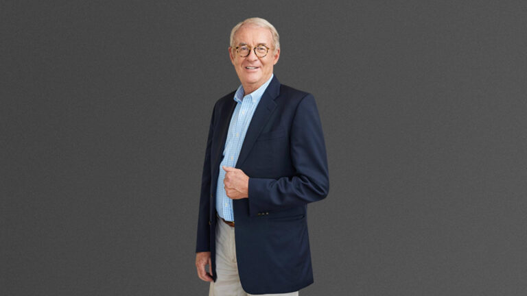 An older man in a dark blazer and light shirt stands against a gray background, smiling softly.