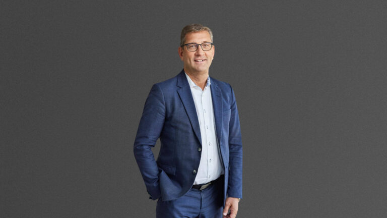 A man in a blue suit and glasses stands confidently against a plain gray background.