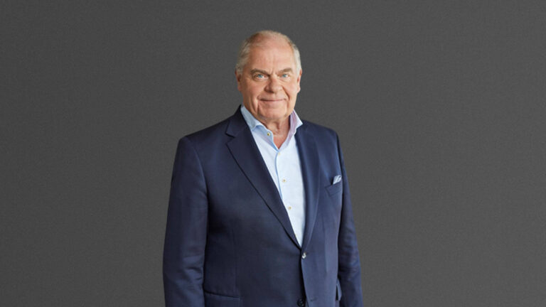 A distinguished older man in a navy suit stands against a plain gray background.