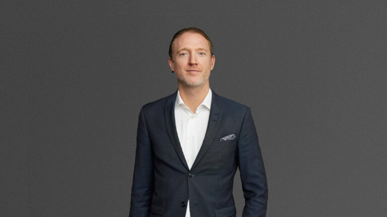 A man standing against a plain, dark background, wearing a dark suit and white shirt.