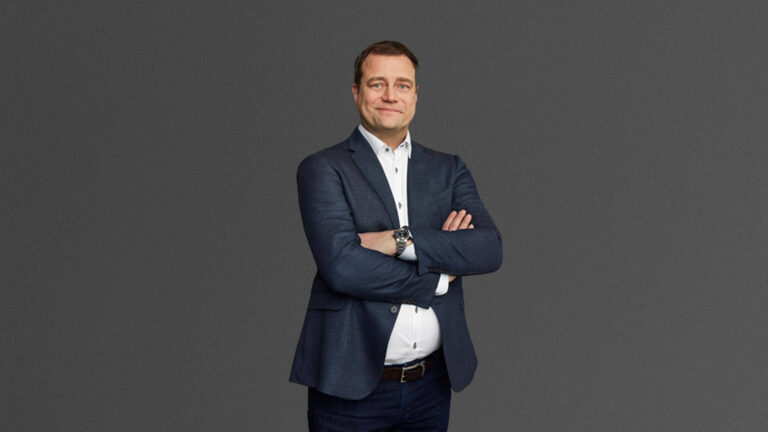 A man in a navy blazer and white shirt stands with arms crossed against a plain gray background.