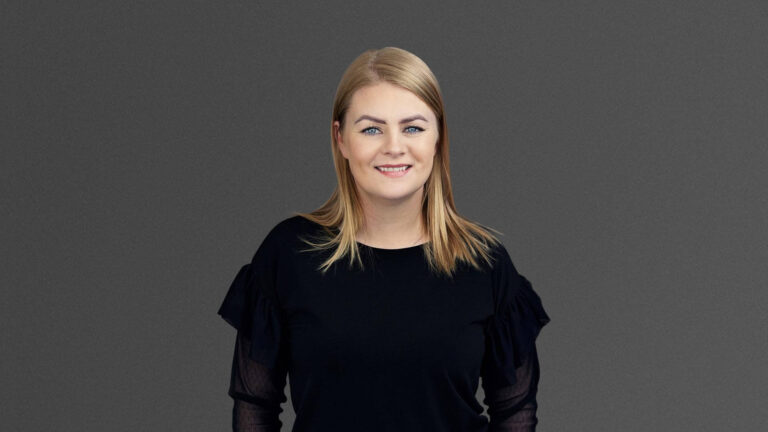 A person with shoulder-length blonde hair, wearing a black top, stands smiling against a gray background.