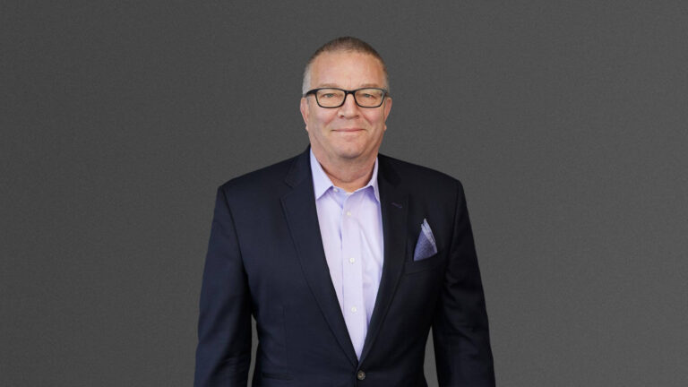 Man in a dark suit and light shirt, wearing glasses, stands against a plain gray background.