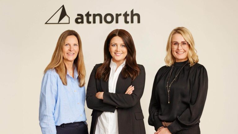 Three women standing in front of an "atnorth" logo, two wearing blazers and one in a blouse.