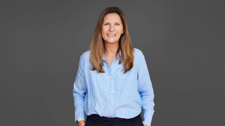 A woman with long hair in a light blue shirt stands against a gray background, smiling with hands in pockets.
