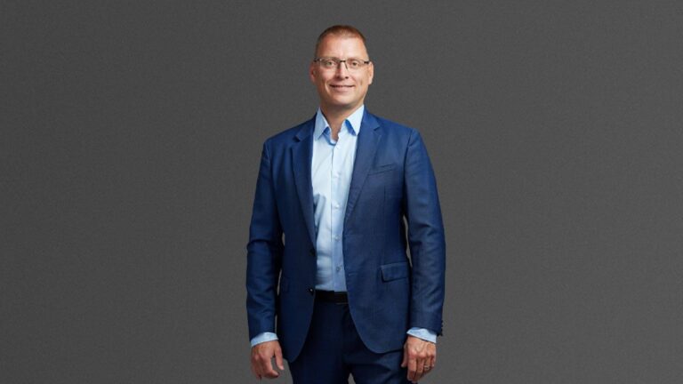 A person in a blue suit and light blue shirt stands against a gray background, facing the camera and smiling.