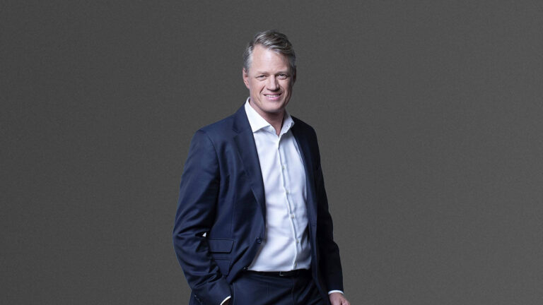 A man in a dark suit and white shirt stands against a plain gray background, smiling with one hand in his pocket.