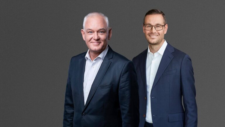 Two men in business suits standing against a plain gray background, both smiling, with one slightly behind the other.