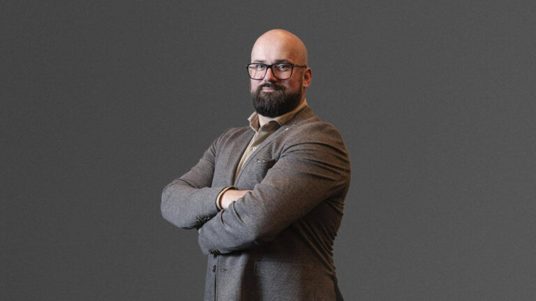 A bearded man with glasses and a bald head stands with arms crossed, wearing a brown blazer against a plain gray background.