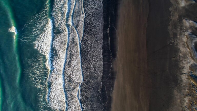 Aerial view of ocean waves breaking onto a dark sandy beach, creating parallel lines of white foam and contrasting textures.