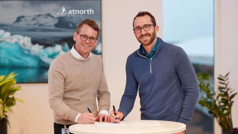 Two men in glasses and sweaters stand smiling and signing papers at a round table. A landscape image and "atnorth" logo are visible in the background.