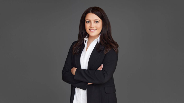 A person with long dark hair wearing a black blazer and white shirt stands confidently against a gray background with arms crossed.