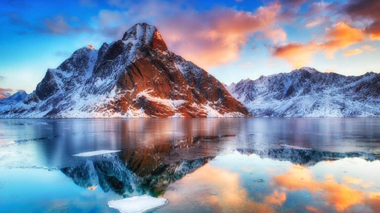 Snow-capped mountains reflected in a serene lake during sunset, with a vibrant sky of orange, pink, and blue hues.