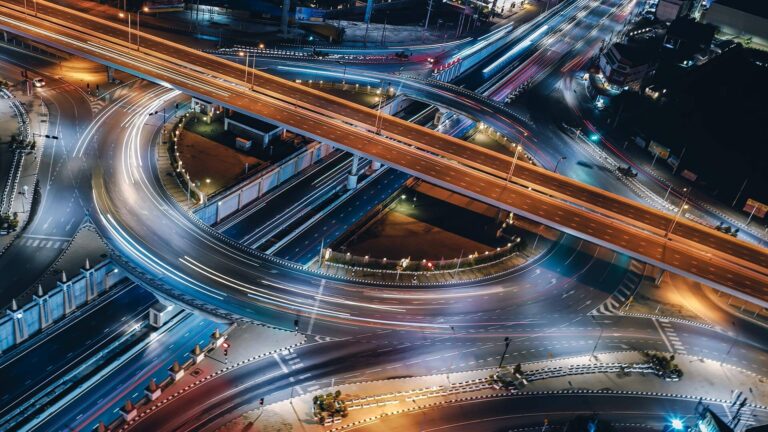 Aerial view of a busy highway interchange at night, with light trails from moving vehicles. Multiple lanes and overpasses create a complex, illuminated network of roads.