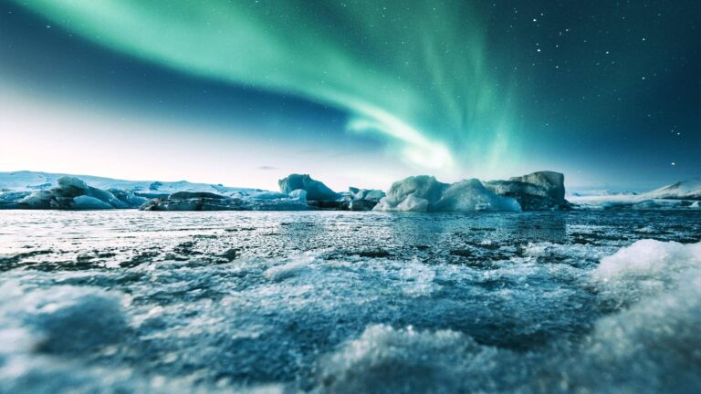 A vibrant green aurora borealis lights up the night sky over a frozen landscape with ice formations and a calm, icy lake.