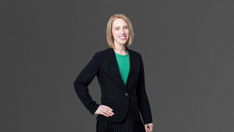 A person with shoulder-length blond hair, wearing a black blazer and green top, stands with one hand on their hip against a gray background.