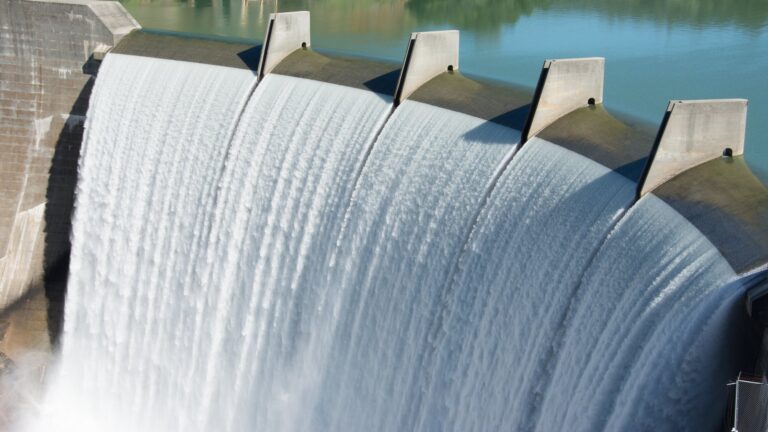 A large dam releases water over its spillway into the reservoir below, with clear blue water reflecting the sky.