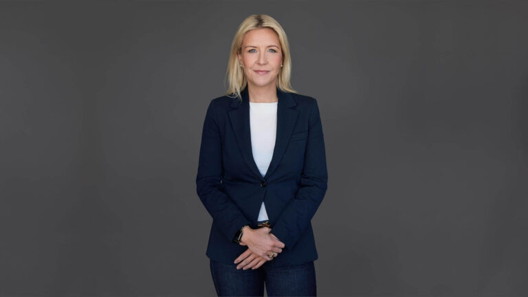 A person with long blonde hair, wearing a dark blazer and jeans, stands against a gray background.