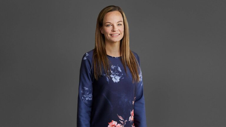 A person with long brown hair wearing a navy shirt with floral patterns stands against a gray background.