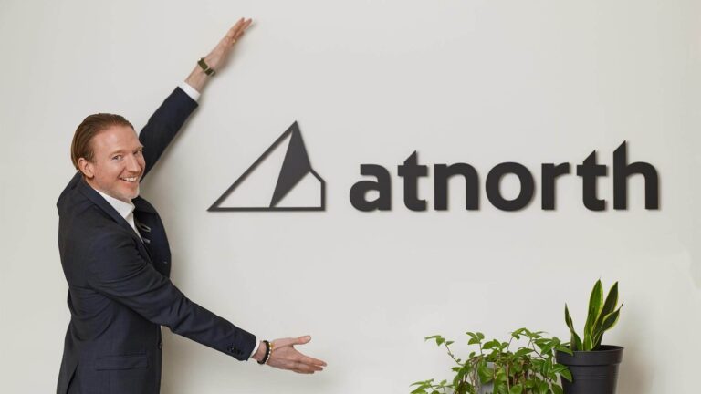 A person in a suit gestures towards a wall with the "atnorth" logo next to a plant.