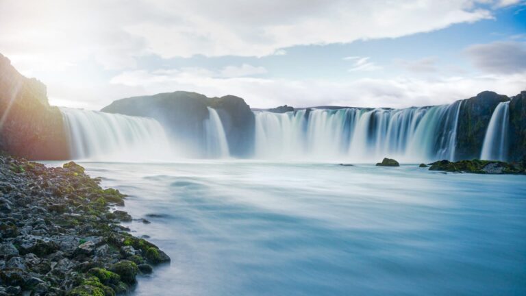Wide-angle view of a waterfall cascading over a rocky cliff into a misty river, with a cloudy sky above.