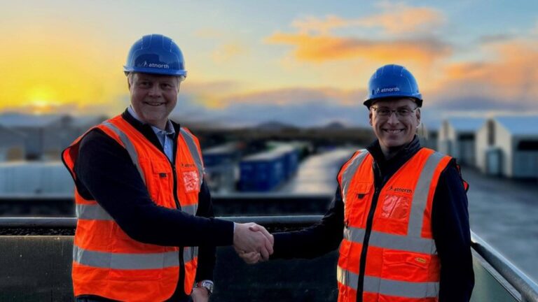 Two people wearing blue hard hats and orange safety vests shake hands at a construction site during sunset.