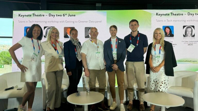 A group of seven people standing together, with event lanyards around their necks, in front of a panel discussion banner.