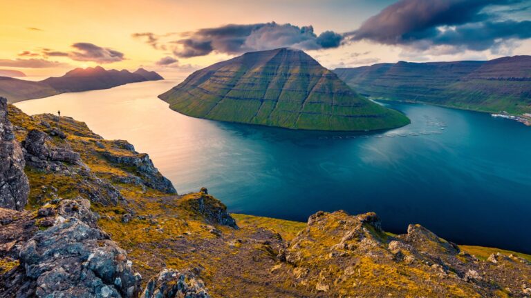 A stunning landscape of a fjord at sunset with steep cliffs and a mountainous island surrounded by calm water.