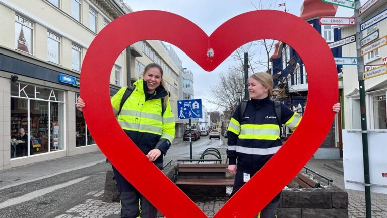 Two people in reflective jackets hold a large red heart frame on a city street, with shops and signs visible in the background.