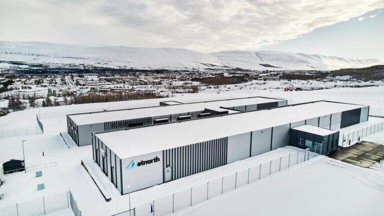 An industrial building covered in snow, surrounded by a fenced area. A snowy landscape and mountains are in the background under a partly cloudy sky.