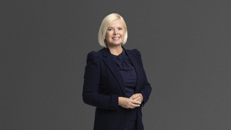 A person with short blonde hair is smiling while wearing a dark suit, standing against a plain gray background.