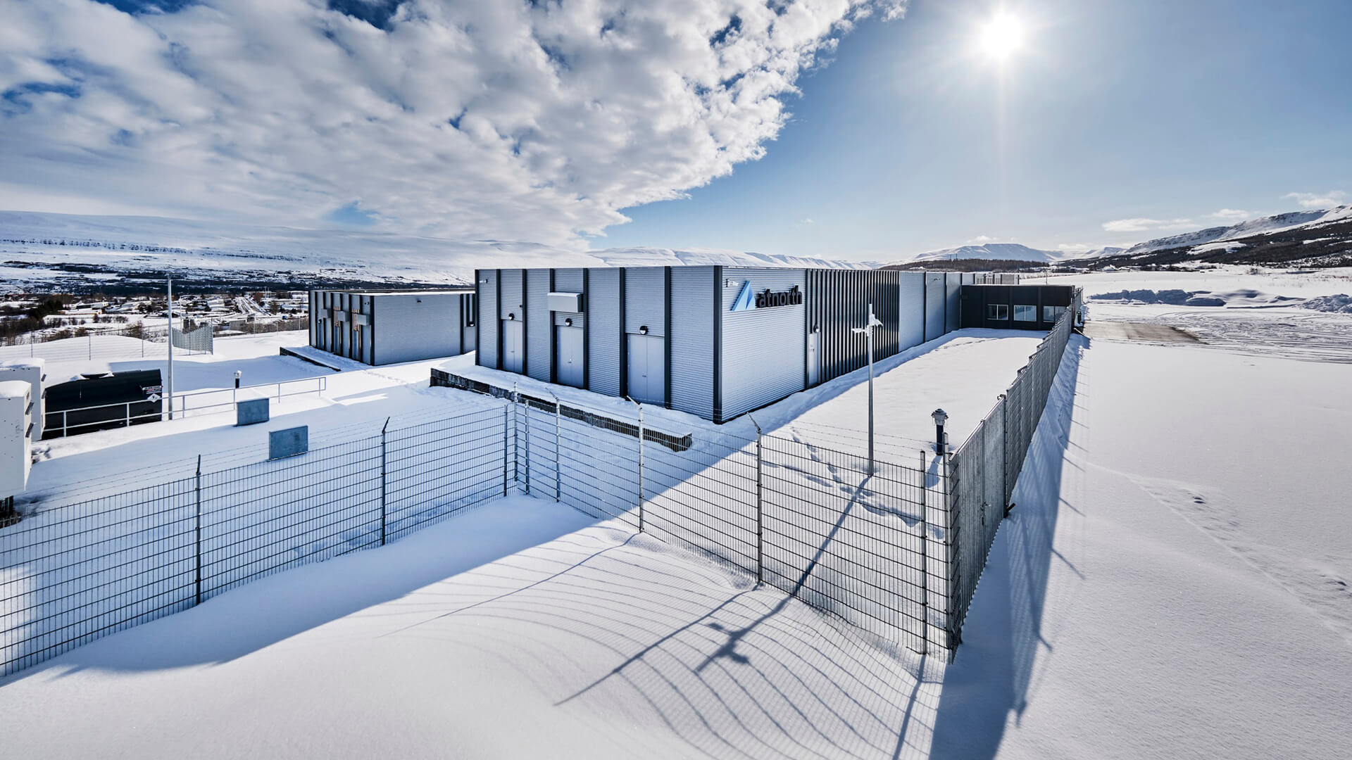 A snow-covered industrial building, possibly housing sustainable data centers, stands with a fenced perimeter under a partly cloudy sky, basking in the sun's glow.