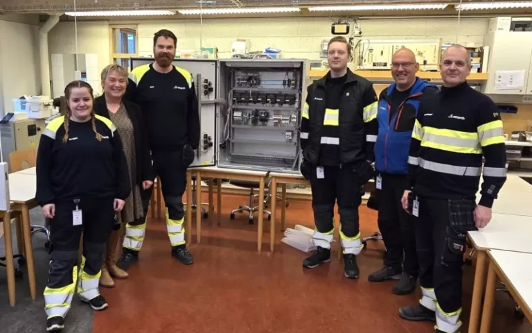 Six people standing in a room with tables, wearing work uniforms and reflective stripes, posing in front of open metal cabinets with electronic equipment.