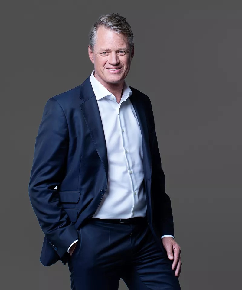 A man in a dark suit with a white shirt smiles with his hand in his pocket against a plain gray background.