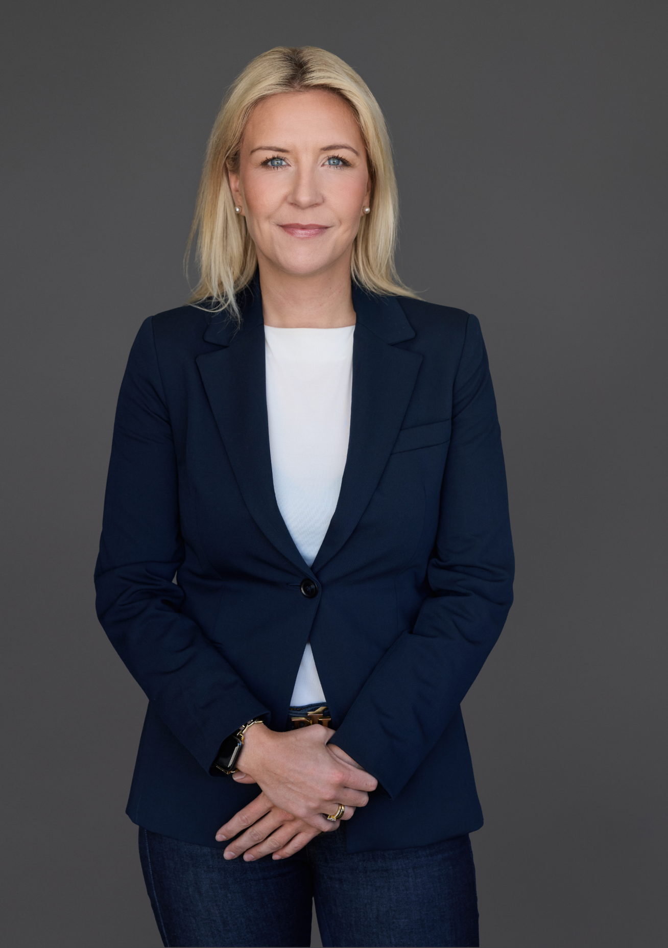 A woman with blonde hair in a navy blazer and white shirt stands confidently against a gray background, embodying the forward-thinking spirit of sustainable data centers, with her hands clasped in front.