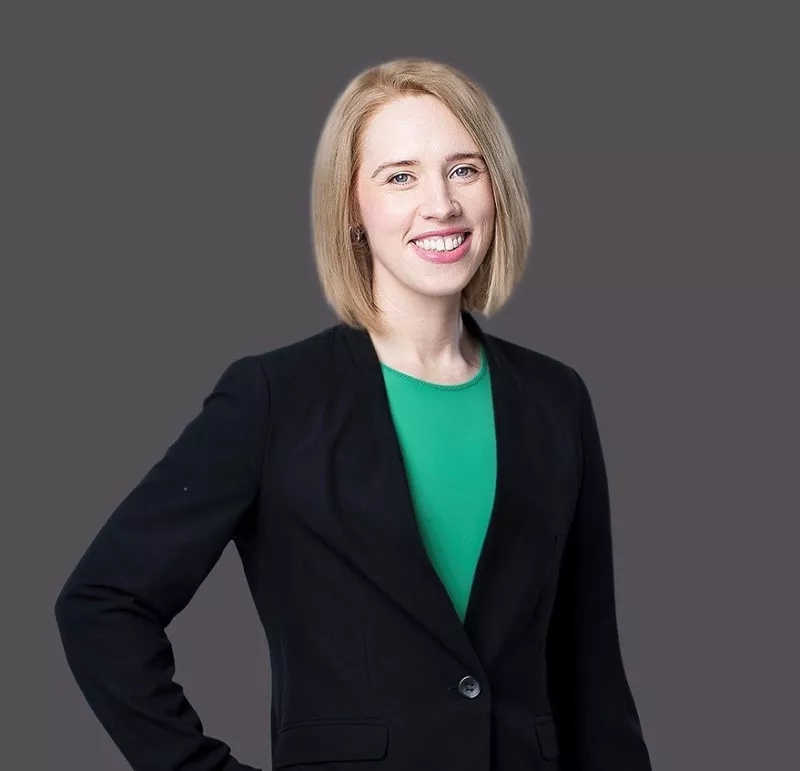 Woman with blonde hair in a black blazer and green top against a gray background, smiling.