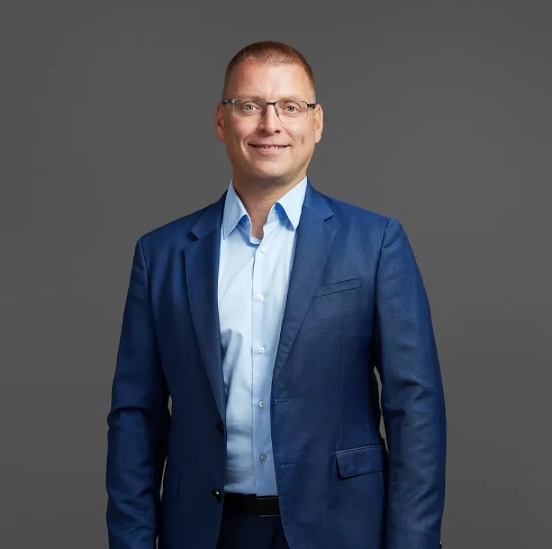 A man in a blue suit and glasses stands against a gray background, smiling.