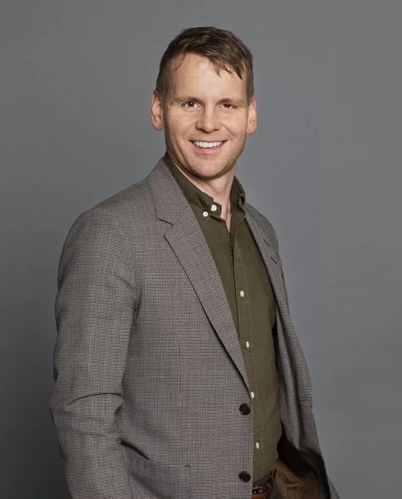 A person in a gray blazer and green shirt smiles against a plain gray background.