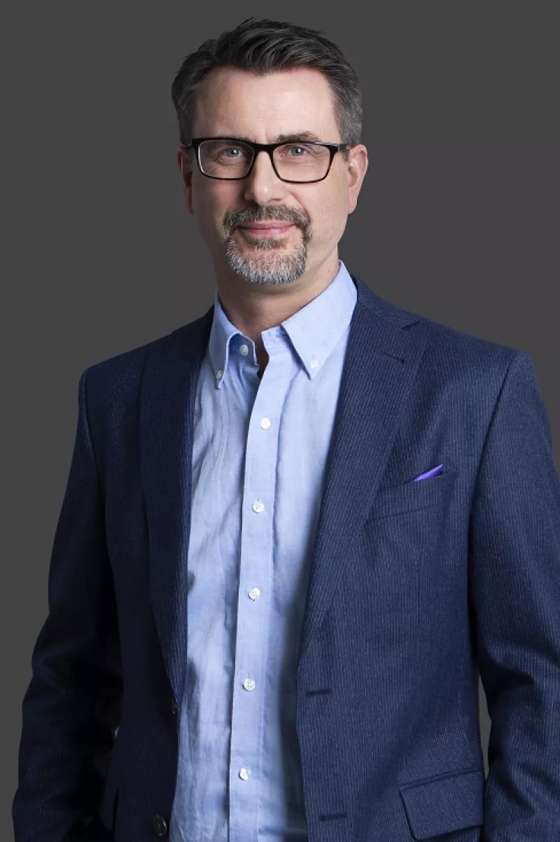 Man in a blue suit and light blue shirt, wearing glasses, stands against a gray background.