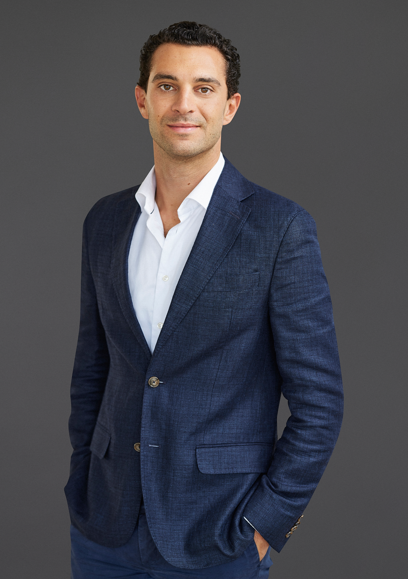 A man in a blue suit jacket and white shirt stands confidently against a gray background, hands in pockets, embodying the sleek professionalism often seen in sustainable data centers.