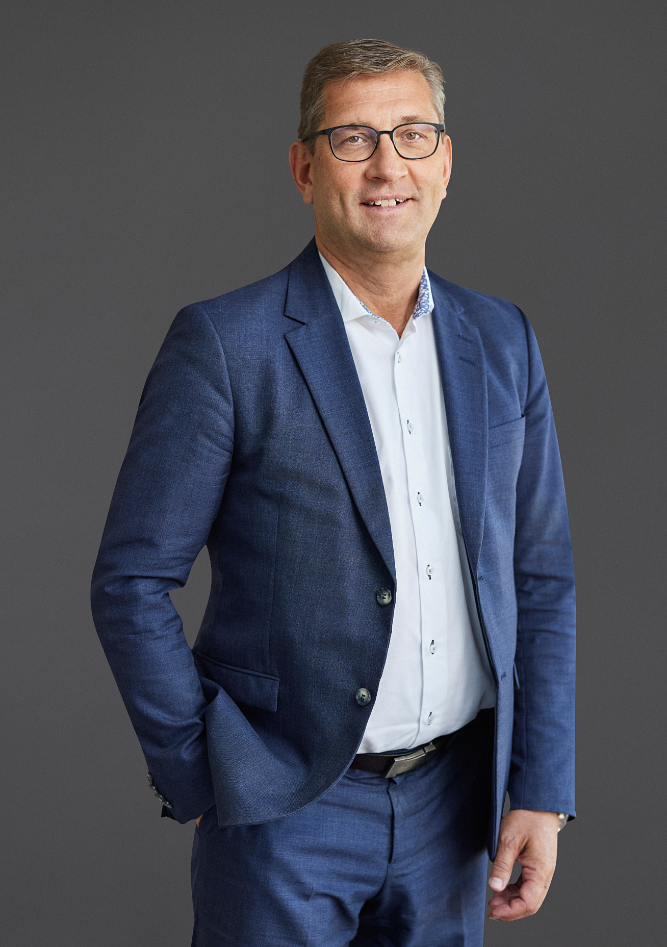 A man in a blue suit and white shirt stands against a gray background, smiling with his left hand in his pocket, embodying the innovation of sustainable data centers.