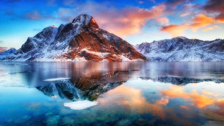 Snow-covered mountains reflected in a calm lake under a vibrant sunset sky, with patches of ice floating on the water's surface.