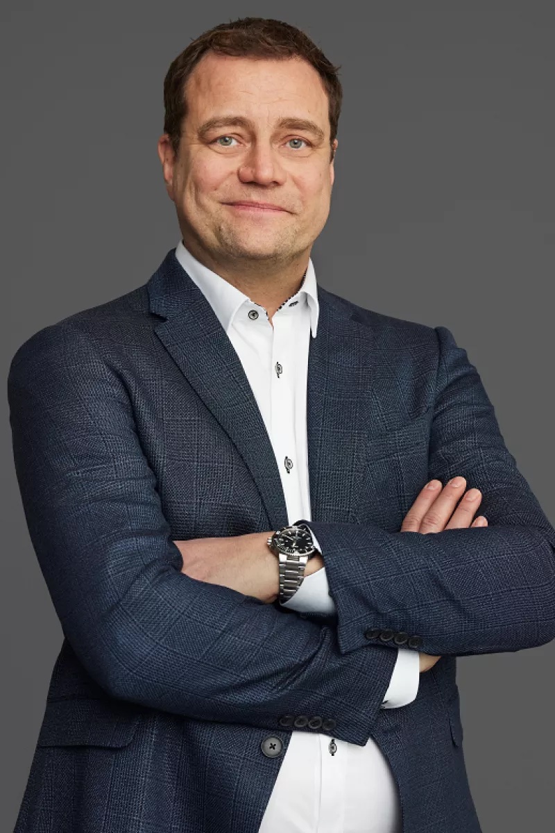 A man in a dark suit and white shirt stands with arms crossed, facing the camera.