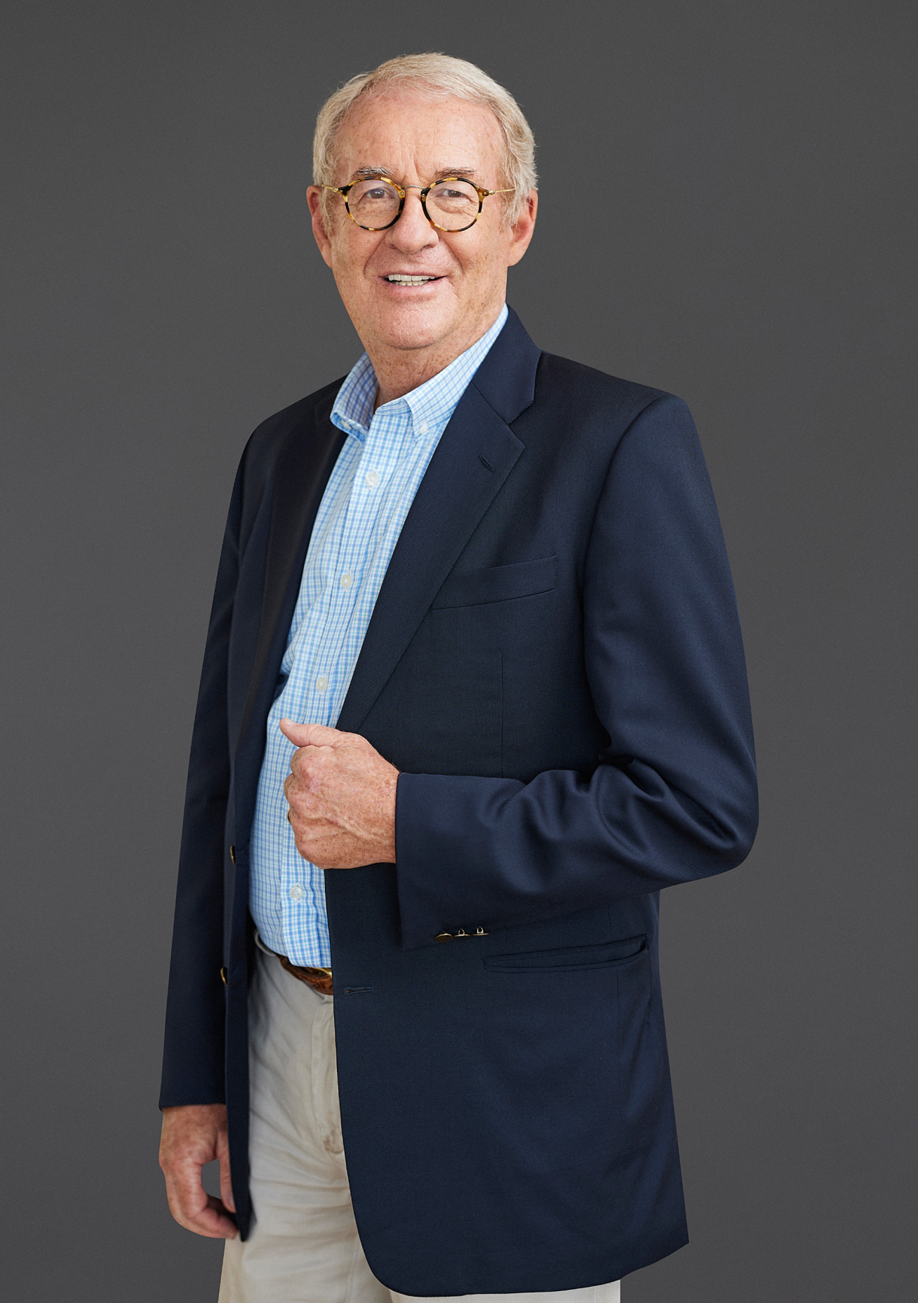 An older man, embodying the principles of sustainable data centers, stands smiling against a gray background, dressed in a navy blazer, light blue shirt, and khaki pants.