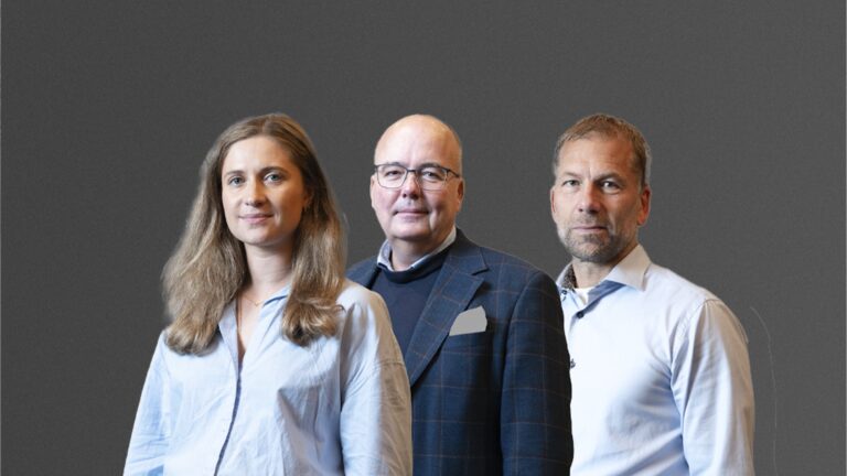 Three people standing in front of a gray background, two men and one woman, all wearing business attire.