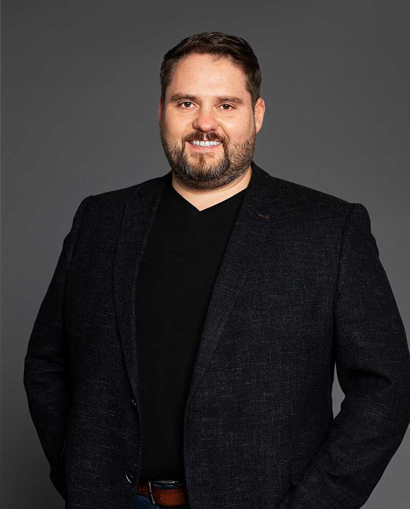 Person wearing a dark blazer and a black shirt, standing against a gray background, smiling at the camera.