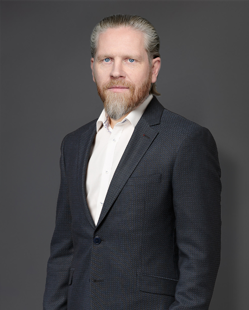 A man with a beard and long hair is wearing a dark suit and white shirt, standing against a gray background.
