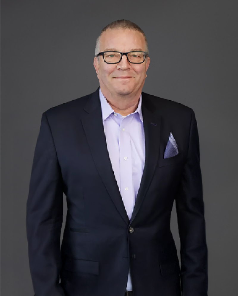 Man in a dark suit and light shirt stands against a gray background, wearing glasses and a slight smile.