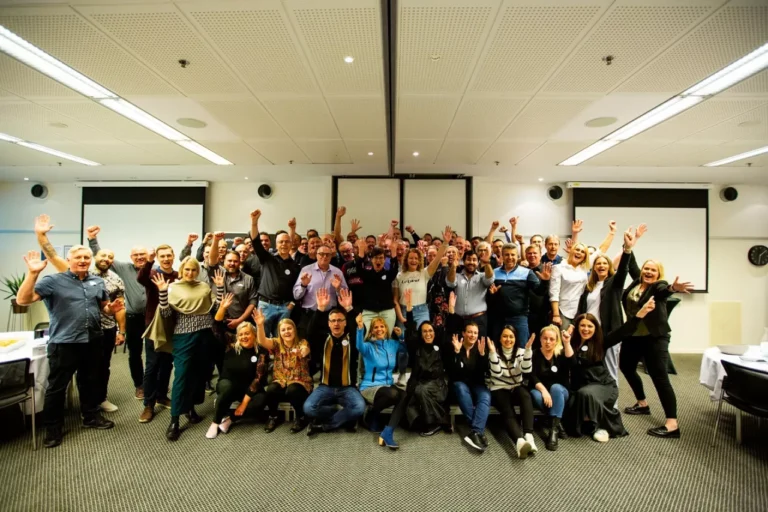 A large group of people posing together in a conference room, with many raising their hands and smiling, is the perfect backdrop as they celebrate the opening of a new data center.