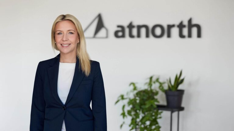 Person in a dark blazer stands smiling in an office setting with "atnorth" logo on the wall and plants in the background.